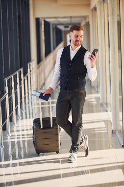 Zarif resmi elbiseli erkek yolcu havaalanı koridorunda bavul, bilet ve telefonla bekliyor..