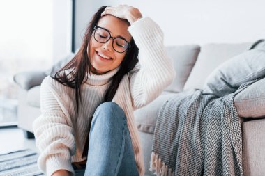 Gündelik giysiler içinde ve gözlüklü genç ve güzel bir kadın evde yalnız oturuyor..