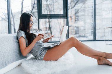 Bir fincan çay ve akıllı telefonla. Seksi, genç ve güzel bir kadın dizüstü bilgisayarla yatağa uzanıyor..