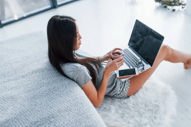 Bir fincan çay ve akıllı telefonla. Seksi, genç ve güzel bir kadın dizüstü bilgisayarla yatağa uzanıyor..