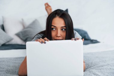 Gündelik giysiler içinde genç ve güzel bir kadın evde dizüstü bilgisayarla yatıyor..