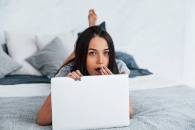 Gündelik giysiler içinde genç ve güzel bir kadın evde dizüstü bilgisayarla yatıyor..