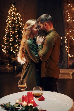 Standing in the Christmas decorated room. Young lovely couple have romantic dinner indoors together.