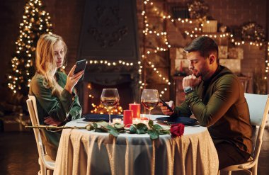 Oturuyor ve telefon kullanıyor. Genç ve güzel çift evde romantik bir akşam yemeği yiyor..