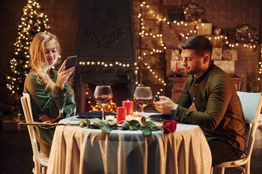 Oturuyor ve telefon kullanıyor. Genç ve güzel çift evde romantik bir akşam yemeği yiyor..