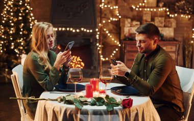 Oturuyor ve telefon kullanıyor. Genç ve güzel çift evde romantik bir akşam yemeği yiyor..