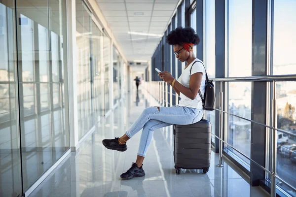 Gündelik giysiler içindeki genç Afro-Amerikan bayan yolcusu havaalanında bavullarıyla bekliyor. Telefon kullanılıyor.