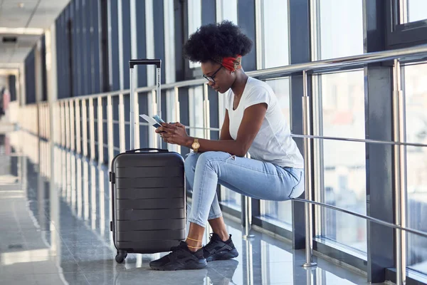 Gündelik giysiler içindeki genç Afro-Amerikan bayan yolcusu havaalanında bavullarıyla bekliyor. Telefon kullanılıyor.