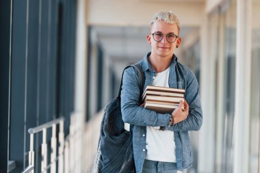 Kot elbiseli genç bir erkek öğrenci, elinde kitaplarla bir üniversitenin koridorunda..