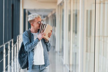 Kot elbiseli genç bir erkek öğrenci, elinde kitaplarla bir üniversitenin koridorunda..