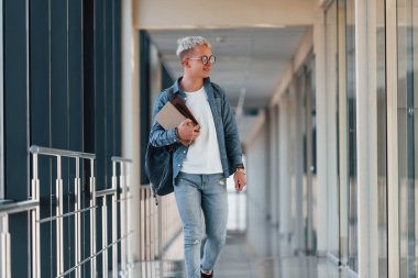 Male young student in jeans clothes is in corridor of a college with books in hands.