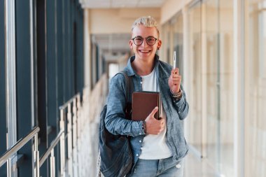 Kot elbiseli genç bir erkek öğrenci, elinde kitaplarla bir üniversitenin koridorunda..