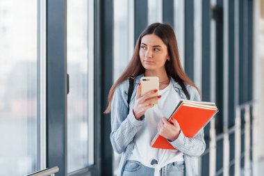 Genç bir kız öğrenci kolejin koridorunda, elinde not defterleri ve telefonlar var..