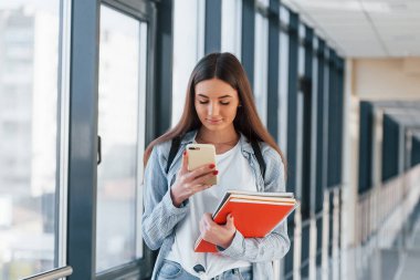 Genç bir kız öğrenci kolejin koridorunda, elinde not defterleri ve telefonlar var..