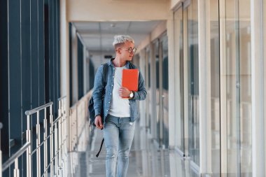 Kot pantolon giymiş genç bir erkek öğrenci elinde not defteriyle bir üniversitenin koridorunda..