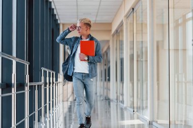 Kot pantolon giymiş genç bir erkek öğrenci elinde not defteriyle bir üniversitenin koridorunda..
