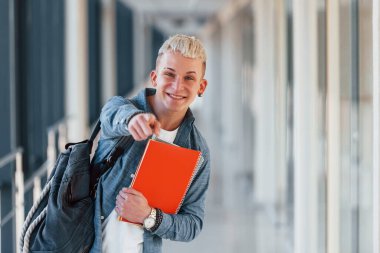 Kot pantolon giymiş genç bir erkek öğrenci elinde not defteriyle bir üniversitenin koridorunda..