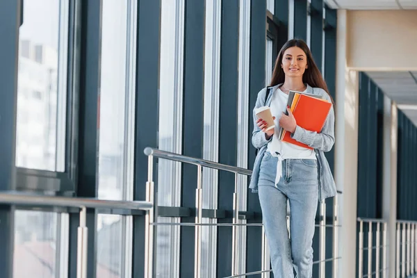 Genç bir kız öğrenci kolejin koridorunda elinde not defterleri ve bir bardak içki tutuyor..