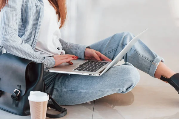 Üniversitenin koridorunda dizüstü bilgisayarı ve yerde bir bardak içkiyle oturan genç kız öğrencinin yakın görüntüsü..