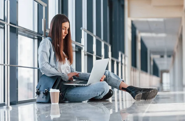 Üniversitenin koridorunda dizüstü bilgisayarı ve yerde bir bardak içkiyle oturan genç bir kız..