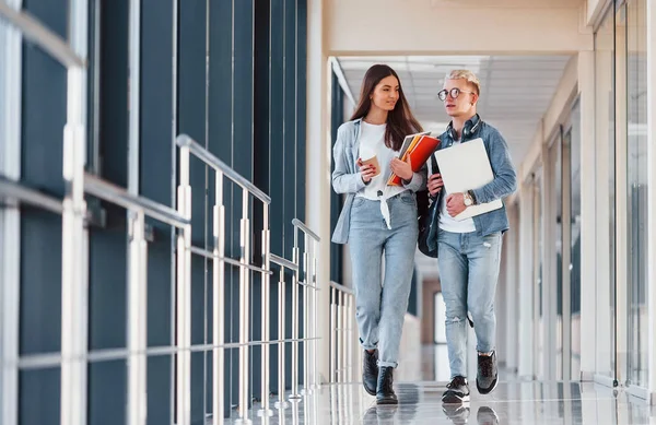 İki genç öğrenci bir kolejin koridorunda birlikte yürüyorlar ve ellerinde not defteri, dizüstü bilgisayar ve bir bardak içki tutuyorlar..