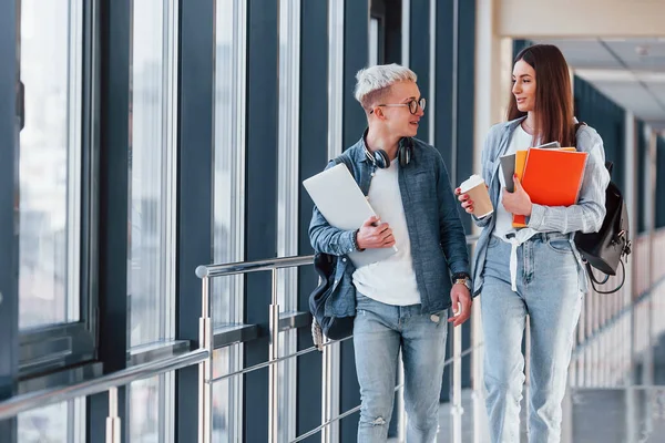 İki genç öğrenci bir kolejin koridorunda birlikte yürüyorlar ve ellerinde not defteri, dizüstü bilgisayar ve bir bardak içki tutuyorlar..