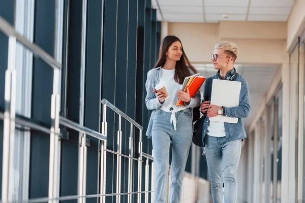 İki genç öğrenci bir kolejin koridorunda birlikte yürüyorlar ve ellerinde not defteri, dizüstü bilgisayar ve bir bardak içki tutuyorlar..