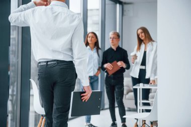 Resmi kıyafetli bir adam yürüyor ve konuşuyor. Ofiste birlikte çalışan ve iletişim kuran başarılı bir grup genç..