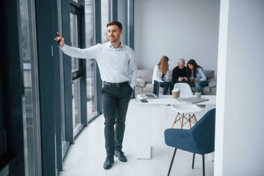 Resmi kıyafetli bir adam, ofiste birlikte çalışan ve iletişim kuran başarılı genç bir takımın önünde duruyor..