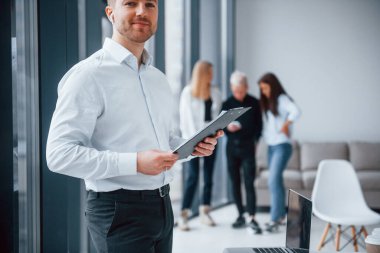 Resmi kıyafetli bir adam, ofiste birlikte çalışan ve iletişim kuran genç ve başarılı bir ekibin önünde duruyor..