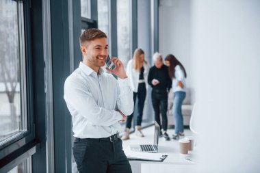 Resmi kıyafetli bir adam telefonla konuşuyor ve ofiste birlikte çalışan ve iletişim kuran başarılı genç bir takımın önünde duruyor..