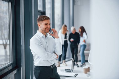 Resmi kıyafetli bir adam telefonla konuşuyor ve ofiste birlikte çalışan ve iletişim kuran başarılı genç bir takımın önünde duruyor..