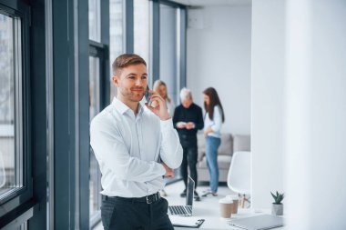 Resmi kıyafetli bir adam telefonla konuşuyor ve ofiste birlikte çalışan ve iletişim kuran başarılı genç bir takımın önünde duruyor..