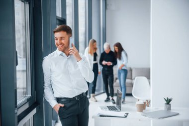 Resmi kıyafetli bir adam telefonla konuşuyor ve ofiste birlikte çalışan ve iletişim kuran başarılı genç bir takımın önünde duruyor..