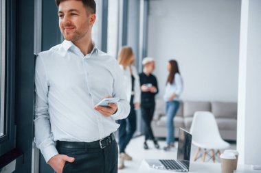 Resmi kıyafetli bir adam elinde telefonla ofiste birlikte çalışan ve iletişim kuran başarılı genç bir takımın önünde duruyor..