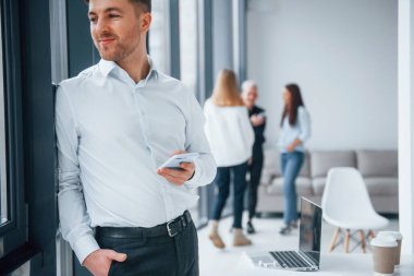 Resmi kıyafetli bir adam elinde telefonla ofiste birlikte çalışan ve iletişim kuran başarılı genç bir takımın önünde duruyor..