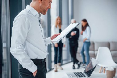 Resmi kıyafetli bir adam, ofiste birlikte çalışan ve iletişim kuran başarılı genç bir takımın önünde duruyor ve belgeleri okuyor..