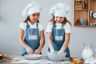 Two little girls in blue chef uniform working with flour on the kitchen.