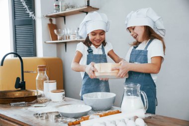 Mavi şef üniformalı iki küçük kız mutfakta elek kullanarak un ile çalışıyorlar..