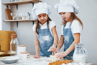 Two little girls in blue chef uniform kneading dough on the kitchen.