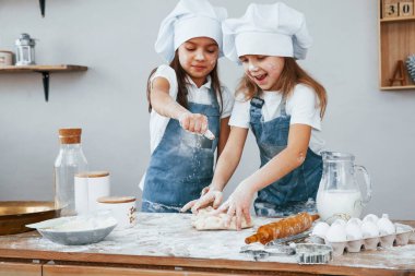 Two little girls in blue chef uniform kneading dough on the kitchen.
