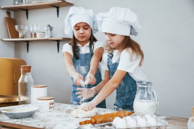 Two little girls in blue chef uniform kneading dough on the kitchen.