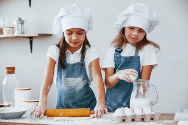 Two little girls in blue chef uniform kneading dough on the kitchen.