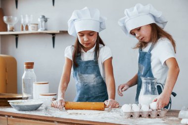 Two little girls in blue chef uniform kneading dough on the kitchen.