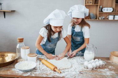 Two little girls in blue chef uniform kneading dough on the kitchen.