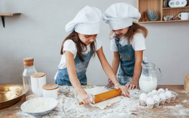 Two little girls in blue chef uniform kneading dough on the kitchen.