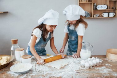 Two little girls in blue chef uniform kneading dough on the kitchen.