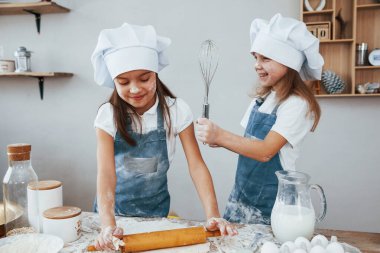 Two little girls in blue chef uniform kneading dough on the kitchen.