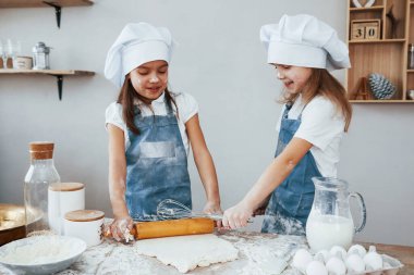 Two little girls in blue chef uniform kneading dough on the kitchen.