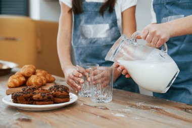 Close up view of two little girls in blue chef uniform that pouring milk into glasses on the kitchen with cookies on table.